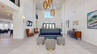 This single-floor community clubhouse is designed with a refined, contemporary aesthetic that immediately impresses upon entry. The grand lobby commands attention with its dramatic double-height ceilings, large transom windows, and statement woven gold pendant lights suspended overhead. Large-format porcelain tile flooring extends throughout, anchoring the open layout with a clean, polished foundation. A central seating arrangement featuring a gray sofa and gold-accented ottomans creates an inviting focal point, while a wall-mounted television and sconce lighting add functional elegance.

Adjacent lounge areas offer additional gathering spaces, including a bright sitting room furnished with terracotta wingback chairs, sheer-draped windows, and a vibrant cactus-themed art piece. A casual dining and lounge area incorporates a built-in banquette with plaid upholstery alongside a dining table, flowing into a well-appointed kitchenette. The kitchen features warm wood cabinetry, white quartz countertops, aqua glass tile backsplash, stainless steel appliances, and a dedicated coffee and beverage station. A separate lounge opens to an outdoor balcony through French doors, filling the room with natural light. Curated artwork and greenery installations throughout complete the polished, welcoming ambiance.