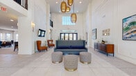 This single-floor community clubhouse is designed with a refined, contemporary aesthetic that immediately impresses upon entry. The grand lobby commands attention with its dramatic double-height ceilings, large transom windows, and statement woven gold pendant lights suspended overhead. Large-format porcelain tile flooring extends throughout, anchoring the open layout with a clean, polished foundation. A central seating arrangement featuring a gray sofa and gold-accented ottomans creates an inviting focal point, while a wall-mounted television and sconce lighting add functional elegance.

Adjacent lounge areas offer additional gathering spaces, including a bright sitting room furnished with terracotta wingback chairs, sheer-draped windows, and a vibrant cactus-themed art piece. A casual dining and lounge area incorporates a built-in banquette with plaid upholstery alongside a dining table, flowing into a well-appointed kitchenette. The kitchen features warm wood cabinetry, white quartz countertops, aqua glass tile backsplash, stainless steel appliances, and a dedicated coffee and beverage station. A separate lounge opens to an outdoor balcony through French doors, filling the room with natural light. Curated artwork and greenery installations throughout complete the polished, welcoming ambiance.