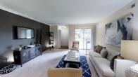 The Elm is a classic 2-bedroom, 2-bathroom single-floor residence that balances comfort and style across a thoughtfully designed floor plan. The living room serves as the heart of the home, featuring plush carpeting, crown molding, and a striking dark accent wall that adds depth and visual contrast to the otherwise light, neutral color palette. A sliding glass door floods the space with natural light while providing views of the outdoors, creating an airy, open atmosphere. The living area comfortably accommodates a full seating arrangement, centered around a glass-top coffee table atop a bold blue patterned area rug, highlighting the room's generous proportions. A built-in shelving unit and media console offer practical storage and display options. The entryway is clearly defined, providing a natural transition into the main living space. With its classic finishes and warm, inviting ambiance, the Elm offers a well-rounded living environment suited for a variety of lifestyles. The combination of natural light, thoughtful design details, and a cohesive color scheme throughout gives this residence a polished, move-in-ready appeal.