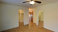 This single-floor space is thoughtfully designed to maximize functionality within a compact layout. The main living area features warm-toned hardwood flooring that extends throughout, creating a cohesive and inviting atmosphere. A ceiling fan with integrated lighting is centrally mounted overhead, providing both air circulation and ambient illumination.

From the main living area, three distinct openings provide access to the space's key zones. To the left, a walk-in closet offers generous storage with visible shelving and hanging rods, making organization straightforward. Directly ahead, the kitchen is open to the living area and is equipped with warm medium-toned wood cabinetry, a white range, and a sink set beneath upper cabinets — offering an efficient and practical cooking space. To the right, a mirrored sliding glass door leads to an additional area, likely a bathroom or secondary closet, with the reflective surface helping to visually expand the space.

The overall aesthetic is clean and neutral, with white walls and trim that complement the natural wood tones throughout, resulting in a bright and functional living environment.