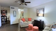 This two-floor space features a welcoming living area with dark hardwood-style flooring that anchors the room with warmth and contrast. A ceiling fan with an integrated light fixture is mounted to the textured ceiling, providing both air circulation and ambient lighting. The living area flows openly toward a connected kitchen, which is visible in the background and equipped with light wood cabinetry, a white refrigerator, and a range. The layout creates an efficient, open feel between the main living and cooking spaces. Neutral-toned walls keep the interior bright and versatile, while decorative wall art adds personality to the space. The living area comfortably accommodates a loveseat accented with coral-toned throw pillows, paired with a dark dresser and ottoman-style seating that introduce color and functional storage. A patterned area rug defines the seating zone on the hardwood-style floor. The overall ambiance is casual and livable, with a practical layout that makes good use of the available space across both floors.