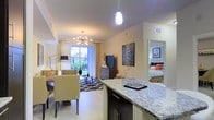 This single-floor residence is designed with an open-concept layout that seamlessly connects the kitchen, dining, and living areas into one cohesive, flowing space. The kitchen features a prominent granite-topped island with dark cabinetry, offering both generous counter space and a natural visual anchor for the open floor plan. A modern pendant light above the island adds a contemporary touch.

The living and dining area is illuminated by a striking sputnik-style chandelier that serves as a focal point, complementing the warm, neutral tones throughout. Large sliding glass doors invite abundant natural light and provide a direct view of lush outdoor greenery, enhancing the sense of openness. Light-toned flooring runs throughout, further contributing to the bright, airy atmosphere.

A patterned area rug grounds the living space, where comfortable seating is arranged around a media console. Decorative wall art adds visual interest and personality to the room. A glimpse into the adjoining bedroom reveals a well-appointed sleeping area with warm accent colors and framed artwork, maintaining the cohesive and inviting aesthetic found throughout the residence.