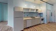 This single-floor loft-style space blends industrial character with contemporary finishes throughout. The open-plan layout is anchored by rustic wood-look flooring that runs continuously across the entire unit, lending warmth and cohesion to every area. Exposed brick walls line one side of the main living area, providing striking textural contrast against the smooth white ceilings and pale blue-painted walls. Industrial-style black pipe conduit runs along the ceiling, reinforcing the loft aesthetic, while a structural support column adds architectural interest to the open floor plan. Tall windows set within the brick wall frame abundant natural light, brightening the generous living area throughout the day. The kitchen is clearly defined along one wall and is fully equipped with a full-size refrigerator, electric range, dishwasher, and a single-basin sink. Light-toned cabinetry with open upper shelving provides ample storage, and a series of pendant lights hang above the countertop, offering both task lighting and a stylish focal point. A panel door adjacent to the kitchen leads to an additional room or closet space. The overall ambiance of this space is bright, airy, and urban, making it well-suited for a variety of living or working arrangements.