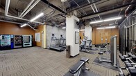 This fitness center delivers a comprehensive workout facility designed to accommodate a variety of exercise routines. The space features an open, industrial-style ceiling with exposed ductwork, pipes, and overhead fluorescent lighting that lends the room a modern, utilitarian aesthetic. Durable, striped carpet tiles cover the flooring throughout, providing a practical and cushioned surface underfoot.

The cardio section is prominently equipped with multiple treadmills, each outfitted with individual display monitors, as well as elliptical machines positioned to allow ample space between users. The strength and functional training area includes a weight bench and additional resistance equipment, offering flexibility for a range of workout programs. A structural column near the center of the room is accented with informational postings, likely workout guides or facility rules.

The warm-toned accent walls contrast with the neutral concrete and exposed infrastructure above, creating an energetic yet grounded atmosphere. A water station is conveniently accessible on the fitness floor. Additional amenities include a vending machine near the entrance and what appears to be a windowed room or storage area along one wall. The layout is open and well-organized, ensuring ease of movement and accessibility throughout the space.