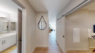 This contemporary single-floor residence offers a thoughtfully designed layout that balances comfort and modern aesthetics. The entryway sets an inviting tone, featuring light wood flooring, crisp neutral walls, a decorative round mirror with a leather strap accent, and a sliding barn door that adds a touch of architectural interest.

The open-concept living area is bathed in abundant natural light streaming through large floor-to-ceiling windows that frame outdoor views. Light wood flooring flows seamlessly throughout, complementing the airy, neutral color palette. The living space offers ample room for relaxed lounging and entertaining.

The kitchen is sleek and functional, featuring grey cabinetry with modern hardware, light stone countertops, stainless steel appliances, and stylish pendant lighting. A kitchen island provides additional workspace and casual seating, while light wood flooring ties the space cohesively to the rest of the home.

The bedroom is a comfortable retreat, highlighted by a textured blue accent wall, large windows that welcome natural light, and generous floor space. The bathroom features clean, contemporary tile work, a glass-enclosed shower stall, and recessed lighting, projecting a polished and efficient design. Together, these spaces create a cohesive, modern living environment with a bright and welcoming ambiance.