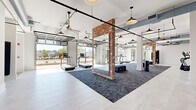 Located at 4271 Woodward Mill Rd in Buford, Georgia, this single-floor fitness center combines industrial-chic design with a bright, welcoming atmosphere. The expansive open-concept layout is anchored by striking exposed brick columns framed with large mirrors, creating a dynamic visual depth throughout the space. Overhead, exposed ductwork and black industrial pipe runs contrast against crisp white ceilings, while a series of pendant lights provide warm, even illumination across the floor.

A standout architectural feature is the row of retractable glass-panel garage doors that open the cardio zone directly to the outdoors, flooding the space with natural light and fresh air. This area houses a long row of modern treadmills and step platforms positioned to take full advantage of the outdoor views. Light-toned tile flooring transitions to textured dark-gray carpet in the central training zones, clearly delineating functional areas.

Additional equipment zones include elliptical trainers, cable resistance machines, and open floor space suited for functional training or group fitness. The entry corridor features a clean, minimal aesthetic with patterned accent wallcovering and a dedicated fitness rules display. Restroom and utility access doors are integrated seamlessly into the layout. The overall ambiance is energetic yet refined, blending modern fitness functionality with thoughtful architectural design.