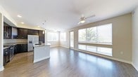 This single-floor residence showcases a thoughtfully designed open-concept layout that seamlessly integrates the kitchen, dining, and living areas into one cohesive, light-filled space. Rich dark hardwood-style flooring runs throughout, providing a striking contrast against the crisp white ceilings and neutral-toned walls.

The modern kitchen is a standout feature, boasting dark cabinetry, a stainless steel refrigerator, and a curved peninsula with pendant lighting above — ideal for casual dining or entertaining. A dedicated sink on the peninsula adds both style and functionality to the space.

The expansive living and dining area is bathed in natural light, courtesy of a large bank of windows fitted with horizontal blinds and a sliding glass door that opens to the outdoors. The generous window coverage provides unobstructed views of mature, lush trees, creating a serene, nature-connected ambiance throughout the day.

A ceiling fan centered in the living area ensures year-round comfort while complementing the clean, contemporary aesthetic of the space. The overall design is airy and welcoming, with an abundance of natural light and an efficient, well-connected floor plan that makes excellent use of every square foot.