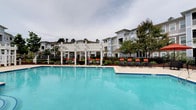 Located at 2222 Crow Ln in Myrtle Beach, South Carolina, this outdoor amenity area offers a well-appointed resort-style retreat designed for relaxation and recreation. The centerpiece of the space is a generously sized swimming pool with clear, blue water and a classic freeform shape, providing ample room for both leisurely swimming and social enjoyment. The pool deck is finished in a light-toned concrete surface and lined with a row of black-framed chaise lounge chairs accented with vibrant orange cushions, creating a cheerful and inviting atmosphere. A standout architectural feature is the white pergola structure positioned at the far end of the pool, offering shaded seating and an elegant focal point to the outdoor layout. Lush landscaping, including palm trees and manicured shrubs, frames the area and adds a tropical coastal ambiance befitting the Myrtle Beach locale. Black wrought-iron fencing encloses the pool area, ensuring both safety and a polished aesthetic. The backdrop features a multi-story residential building clad in soft blue-gray siding with balconies overlooking the pool, further enhancing the sense of community and cohesion throughout the property.