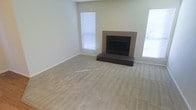 This single-story space centers around a spacious living area finished with wall-to-wall neutral-toned carpet, creating a warm and understated foundation throughout. The room's most prominent feature is a tile-surround fireplace positioned along one wall, complete with a raised hearth finished in dark tile, adding a classic focal point to the space. The fireplace surround's earthy tones complement the light, neutral wall color, contributing to a cohesive and understated aesthetic. Natural light enters generously through multiple windows fitted with horizontal blinds, allowing for adjustable brightness and privacy. The walls are painted in a clean, soft off-white tone that enhances the sense of openness. White baseboards frame the perimeter of the room, providing a crisp contrast against the carpeted floor. The overall layout offers a generous, open feel with room to configure furnishings in a variety of arrangements. The space presents a clean, move-in ready condition, with a straightforward and functional design well-suited for comfortable everyday living.