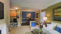 This single-floor residence showcases an open-concept layout that seamlessly connects the living, dining, and kitchen areas, creating a cohesive and functional flow throughout the space. The living area features plush carpeting and is furnished with neutral-toned seating accented by vibrant blue throw pillows, adding a pop of color against the warm olive-green accent wall. A glass-top coffee table anchors the seating arrangement, while a side table with a classic lamp adds ambient lighting to the corner.

The dining area is defined by a striking navy-blue accent wall adorned with framed artwork, creating a bold focal point. A pendant light fixture hangs above the dark dining table and chairs, providing both task lighting and visual interest. The transition between spaces is marked by a change from carpet to dark hardwood-style flooring, adding architectural definition.

The kitchen is partially visible beyond a raised breakfast bar with dark bar stools, offering casual seating alongside the formal dining space. Light wood cabinetry and under-cabinet lighting contribute warmth to the kitchen area. Throughout the space, the combination of earthy tones, bold accents, and layered lighting creates an inviting and well-appointed atmosphere.