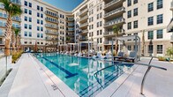 Located at 180 Patriots Point Rd in Mount Pleasant, South Carolina, this impressive outdoor amenity space is set within a beautifully designed courtyard enclosed by a multi-story residential building with classic Charleston-inspired architecture. The centerpiece is a generously sized resort-style swimming pool with crystal-clear blue water, flanked by wide pool decks finished in light-toned pavers. Sleek lounge chairs, modern pergola-style cabanas, and lush palm trees line the pool's perimeter, creating a refined, resort-like atmosphere.

Adjacent to the pool, a covered pergola lounge area features a slatted metal overhead structure strung with bistro lighting, exposed brick accent walls, and vibrant teal bar-height seating arranged around a counter — ideal for casual outdoor gatherings. Lush tropical plantings add natural color and texture to this zone.

A separate courtyard terrace showcases a striking geometric patterned tile floor in shades of grey and white, furnished with contemporary lounge chairs and sofas in neutral tones accented with teal throw pillows. A manicured grass lawn at the center of this area provides a verdant focal point. Glass railings and architectural pergola elements tie the spaces together, while the surrounding building's balconies and large windows frame the entire courtyard with an elegant, layered backdrop.