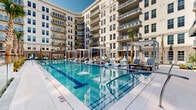 Located at 180 Patriots Point Rd in Mount Pleasant, South Carolina, this impressive outdoor amenity space is set within a beautifully designed courtyard enclosed by a multi-story residential building with classic Charleston-inspired architecture. The centerpiece is a generously sized resort-style swimming pool with crystal-clear blue water, flanked by wide pool decks finished in light-toned pavers. Sleek lounge chairs, modern pergola-style cabanas, and lush palm trees line the pool's perimeter, creating a refined, resort-like atmosphere.

Adjacent to the pool, a covered pergola lounge area features a slatted metal overhead structure strung with bistro lighting, exposed brick accent walls, and vibrant teal bar-height seating arranged around a counter — ideal for casual outdoor gatherings. Lush tropical plantings add natural color and texture to this zone.

A separate courtyard terrace showcases a striking geometric patterned tile floor in shades of grey and white, furnished with contemporary lounge chairs and sofas in neutral tones accented with teal throw pillows. A manicured grass lawn at the center of this area provides a verdant focal point. Glass railings and architectural pergola elements tie the spaces together, while the surrounding building's balconies and large windows frame the entire courtyard with an elegant, layered backdrop.