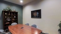 This single-floor professional space is thoughtfully designed to accommodate meetings and collaborative gatherings in a polished, focused environment. The centerpiece of the room is a rich, warm-toned wooden conference table surrounded by sleek gray upholstered chairs, offering both comfort and a refined aesthetic. The walls are finished in a soft neutral tone, complemented by an accent wall in a muted sage green that adds depth and a calming visual contrast to the space. A framed vintage-style world map with an ornate dark border serves as a striking decorative focal point on the main wall, lending the room a sophisticated, worldly character. To one side, a dark wood bookshelf displays curated decorative objects, adding a layer of warmth and personality to the otherwise clean, professional interior. Recessed lighting illuminates the space evenly from above, creating a well-lit atmosphere suitable for productive work sessions. The overall ambiance is professional yet inviting, striking a balance between functionality and tasteful design — making it well-suited for client meetings, team discussions, or focused work.
