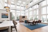 This exceptional amenity space is defined by its dramatic double-height ceilings and an expansive curtain wall of floor-to-ceiling windows that flood the interior with natural light and frame sweeping views of surrounding treetops and the cityscape beyond. Warm hardwood flooring flows throughout, anchoring the open-plan layout that seamlessly connects multiple distinct zones. The lounge area features a generous sectional sofa, a sculptural teal curved couch, and curated accent chairs arranged around a marble-top coffee table, all set atop vibrant area rugs in teal and rose tones. A long dining table with leather chairs provides a formal gathering space, while a slatted wood media wall adds warmth and visual structure. The fully equipped kitchen showcases a large marble-topped island with bar seating, sleek cabinetry, marble-look tile backsplash with under-lit open shelving, and stainless steel double wall ovens. A stylish banquette dining nook is highlighted by a striking large-format teal and gold abstract mural, complemented by gold-accented furniture and adjustable wall sconces. Thoughtful details throughout include designer pendant lighting, a water station, and a kitchenette wet bar, all contributing to a polished, sophisticated, and welcoming atmosphere.