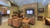 This single-floor space offers a warm and inviting lounge environment designed for comfort and gathering. The focal point of the room is a dramatic arched alcove finished in stacked stone, which frames a large flat-screen television and serves as a commanding architectural centerpiece. The stacked stone detail adds texture and a sense of craftsmanship, complementing the room's earthy, neutral color palette of warm tans and beiges.

The seating area is thoughtfully arranged to encourage conversation and relaxation, featuring striped upholstered armchairs, a rich burgundy sofa, and wooden coffee tables that anchor the space. Recessed lighting in the ceiling provides consistent, even illumination throughout, enhancing the room's welcoming ambiance.

Decorative touches such as framed artwork, ornamental wall accents, and potted indoor plants add personality and a lived-in warmth to the environment. The flooring is carpeted in a neutral tone that ties the color scheme together seamlessly.

The layout is open and spacious, allowing for comfortable movement between seating clusters. The overall design aesthetic blends transitional and Mediterranean influences, creating a timeless and approachable atmosphere well-suited for lounging, socializing, or enjoying media in a relaxed communal setting.