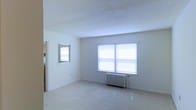 This single-floor space offers a clean and straightforward layout that provides a solid foundation for comfortable living. The interior is characterized by crisp white walls and a textured ceiling that lends a classic residential feel throughout. The main living area is generously proportioned, bathed in natural light streaming through multiple windows fitted with horizontal blinds, creating a bright and airy atmosphere. A traditional cast-iron radiator beneath the windows serves as a notable architectural detail, reflecting the building's classic heating infrastructure while adding a touch of vintage character to the room. The light-toned flooring reflects the abundant natural light, further enhancing the sense of openness. A smaller window on the adjacent wall provides additional ventilation and light. An open doorway connects the main living area to additional spaces within the home, suggesting a practical and accessible floor plan. The overall ambiance is calm, neutral, and move-in ready, offering a versatile blank canvas well-suited for a variety of interior styles and furnishing arrangements.