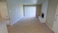 This single-floor space features a spacious living area designed with simplicity and functionality in mind. The room is finished with wall-to-wall beige carpeting that adds warmth underfoot and complements the neutral, light-colored walls throughout. A textured popcorn ceiling adds a classic architectural detail overhead.

A standout feature of the living area is the brick fireplace situated in the corner, finished in white-painted brick and equipped with a black firebox insert. It serves as a focal point that adds character to the otherwise understated space. Adjacent to the fireplace, a built-in white cabinet or shelving unit provides additional storage or display potential.

Natural light is introduced through a large sliding glass door dressed with vertical white blinds, which span nearly the full width of the wall and offer both privacy and light control. The door likely leads to an outdoor area, further enhancing the sense of openness in the room.

Overall, the space presents a clean, neutral palette with practical finishes, making it a versatile and move-in ready environment suitable for a variety of furnishing styles and personal tastes.