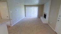 This single-floor space features a spacious living area designed with simplicity and functionality in mind. The room is finished with wall-to-wall beige carpeting that adds warmth underfoot and complements the neutral, light-colored walls throughout. A textured popcorn ceiling adds a classic architectural detail overhead.

A standout feature of the living area is the brick fireplace situated in the corner, finished in white-painted brick and equipped with a black firebox insert. It serves as a focal point that adds character to the otherwise understated space. Adjacent to the fireplace, a built-in white cabinet or shelving unit provides additional storage or display potential.

Natural light is introduced through a large sliding glass door dressed with vertical white blinds, which span nearly the full width of the wall and offer both privacy and light control. The door likely leads to an outdoor area, further enhancing the sense of openness in the room.

Overall, the space presents a clean, neutral palette with practical finishes, making it a versatile and move-in ready environment suitable for a variety of furnishing styles and personal tastes.