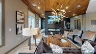 This thoughtfully designed single-floor amenity space blends rustic warmth with contemporary sophistication throughout its open, flowing layout. The centerpiece of the lounge area is a striking reclaimed wood-plank ceiling that adds organic texture overhead, complemented by a dramatic sculptural chandelier with starburst branches that casts ambient light across the room. Large windows line the perimeter, flooding the space with natural light and offering views of the surrounding landscape.

A bold, dark-paneled column anchors the lounge, housing a wall-mounted display above a sleek linear fireplace, creating a natural focal point around which plush seating arrangements are thoughtfully arranged atop layered area rugs over large-format stone tile flooring. A billiards table occupies its own defined zone, adding a recreational dimension to the space.

The kitchen bar area offers a clean, modern aesthetic with a quartz-topped island edged in geometric tile, globe pendant lighting, a textured mosaic backsplash, and a curated arrangement of circular gold-framed mirrors adorning the wall. Appliances and ample counter space make this area well-suited for entertaining.

A private conference room, accessible via a barn-style sliding door, features a deep navy accent wall, a large dining table with upholstered seating, and the same reclaimed wood ceiling, maintaining design continuity throughout the space.