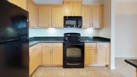 This single-floor residence offers a well-organized layout with a cohesive neutral palette throughout. The kitchen is a functional highlight, featuring light maple cabinetry with brushed nickel hardware, dark green granite countertops, and a full suite of black appliances including a refrigerator, electric range, and built-in microwave. Tile flooring runs through the kitchen, transitioning to plush wall-to-wall carpeting in the living and bedroom areas.

The spacious living area is defined by crown molding, a decorative arched alcove, and a ceiling fan with light fixture. A sliding glass door floods the room with natural light and opens to a balcony with outdoor views. A secondary carpeted bedroom includes a walk-in closet with built-in shelving and a nearby additional closet for ample storage.

The bathroom is notably accessibility-focused, equipped with a roll-in shower featuring multiple grab bars, a handheld showerhead, and a recessed ceiling light. A separate vanity area includes a wide mirror, warm overhead lighting, a single-basin sink, and light maple cabinetry. Tan ceramic tile floors complete the bathroom. Overall, the space maintains a clean, functional, and welcoming ambiance throughout.