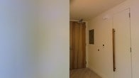 This single-floor space presents a minimalist and contemporary design aesthetic, as evidenced by its clean lines and neutral color palette. The entryway is defined by a solid wood door with warm, natural grain tones, fitted with a door closer mechanism that adds a functional, commercial-grade touch. The walls are finished in a crisp off-white tone, contributing to a bright and airy atmosphere. Light hardwood flooring runs through the entry area, adding warmth to the otherwise understated palette. A recessed panel, likely an electrical or utility access point, is mounted flush to the wall, maintaining the clean visual continuity of the space. Additional wall-mounted controls nearby suggest modern smart or integrated building systems. The ceiling is smooth and unadorned, reinforcing the space's streamlined character. Overall, the design prioritizes functionality and simplicity, with carefully selected materials that balance warmth and modernity. The space appears well-maintained and move-in ready, offering a neutral foundation adaptable to a variety of uses.