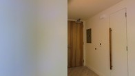 This single-floor space presents a minimalist and contemporary design aesthetic, as evidenced by its clean lines and neutral color palette. The entryway is defined by a solid wood door with warm, natural grain tones, fitted with a door closer mechanism that adds a functional, commercial-grade touch. The walls are finished in a crisp off-white tone, contributing to a bright and airy atmosphere. Light hardwood flooring runs through the entry area, adding warmth to the otherwise understated palette. A recessed panel, likely an electrical or utility access point, is mounted flush to the wall, maintaining the clean visual continuity of the space. Additional wall-mounted controls nearby suggest modern smart or integrated building systems. The ceiling is smooth and unadorned, reinforcing the space's streamlined character. Overall, the design prioritizes functionality and simplicity, with carefully selected materials that balance warmth and modernity. The space appears well-maintained and move-in ready, offering a neutral foundation adaptable to a variety of uses.