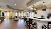 This single-floor space presents an open-concept design that seamlessly blends a social lounge area with a bar-style counter station. The flooring throughout consists of large-format dark tile, lending a clean and contemporary feel to the expansive interior. A tray ceiling with recessed lighting defines the lounge area, adding architectural depth and a sense of elevated elegance. A prominent drum-style pendant light anchors the bar counter zone, complemented by a pair of dark cylindrical pendant lights that hang above the white quartz countertop. The bar counter features high-top seating with dark wood barstools upholstered in bold patterned fabric, and the backdrop is finished with a striking stacked stone accent wall that adds texture and visual interest. The lounge area is furnished with neutral-toned sofas and armchairs arranged around a glass-top coffee table, creating a relaxed and welcoming gathering space. Large windows dressed with sheer and solid curtains allow ample natural light to fill the room. A bold yellow abstract artwork punctuates one wall, adding a vibrant pop of color. The overall ambiance is warm, polished, and well-appointed, making it well-suited for both casual socializing and community gatherings.