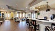This single-floor space presents an open-concept design that seamlessly blends a social lounge area with a bar-style counter station. The flooring throughout consists of large-format dark tile, lending a clean and contemporary feel to the expansive interior. A tray ceiling with recessed lighting defines the lounge area, adding architectural depth and a sense of elevated elegance. A prominent drum-style pendant light anchors the bar counter zone, complemented by a pair of dark cylindrical pendant lights that hang above the white quartz countertop. The bar counter features high-top seating with dark wood barstools upholstered in bold patterned fabric, and the backdrop is finished with a striking stacked stone accent wall that adds texture and visual interest. The lounge area is furnished with neutral-toned sofas and armchairs arranged around a glass-top coffee table, creating a relaxed and welcoming gathering space. Large windows dressed with sheer and solid curtains allow ample natural light to fill the room. A bold yellow abstract artwork punctuates one wall, adding a vibrant pop of color. The overall ambiance is warm, polished, and well-appointed, making it well-suited for both casual socializing and community gatherings.