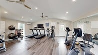 Located at 1587 Briarfield Rd in Hampton, VA, this dedicated fitness center offers a thoughtfully organized exercise space and a supporting bathroom across a single floor. The exercise room features vaulted ceilings that lend an open, airy feel, complemented by recessed lighting and a ceiling fan for comfortable year-round use. A wall-mounted split air conditioning unit provides additional climate control. The flooring throughout is durable wood-look vinyl plank, lending a clean and modern aesthetic. Large windows along the walls allow natural light to fill the space and provide views of the surrounding outdoor area. The fitness area is well-equipped with multiple treadmills, an elliptical trainer, a stationary upright bike, a recumbent bike, a multi-station cable machine, an adjustable weight bench, medicine balls, and a full dumbbell rack. A wall-mounted flat-screen television and a large mirror along one wall enhance the workout environment. The accessible bathroom is finished in the same neutral palette, featuring wood-look vinyl plank flooring, a toilet with grab bars, a wall-mounted sink, and a hand dryer, making it functional and ADA-considerate. Overall, this fitness center provides a bright, clean, and fully equipped space suitable for a well-rounded exercise routine.
