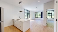 Located at 248 Tioronda Avenue in Beacon, NY, this thoughtfully designed one-bedroom, one-bathroom residence offers a modern aesthetic with high-quality finishes throughout its single-floor layout. The open-concept kitchen and living area serves as the heart of the home, anchored by a sleek white peninsula with a quartz countertop and an undermount sink with a matte black faucet. The kitchen is equipped with stainless steel appliances, including a refrigerator, range, and over-the-range microwave, complemented by white flat-panel cabinetry and a linear pendant light fixture overhead. Light wood-look tile flooring flows seamlessly throughout the entire space, adding warmth and continuity. Oversized grid-pane windows with bold black frames flood both the living area and bedroom with abundant natural light and offer lush greenery views. The generously sized bedroom features a double-door closet and a circular overhead light fixture, with additional closet storage available elsewhere in the unit. The full bathroom is finished with neutral large-format wall tiles, a soaking tub with shower, a wood-toned vanity, and a backlit LED mirror. A dedicated laundry area completes this well-appointed residence.