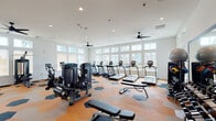 This fitness center in Slidell, Louisiana offers a bright, modern workout environment designed to accommodate a wide range of exercise routines. The single-floor layout is thoughtfully organized, providing ample space for both cardio and strength training activities. A full wall of large, floor-to-ceiling windows lines one side of the facility, flooding the interior with natural light and offering views of the surrounding community, including a pool area and landscaped grounds. The open floor plan is anchored by a distinctive hexagonal-patterned carpet in warm orange and navy tones, giving the space a contemporary, energetic feel. Multiple ceiling fans are installed throughout, complementing the recessed lighting above to maintain a comfortable atmosphere during workouts. The cardio section features a row of True-brand treadmills and elliptical machines positioned along the windowed wall, while spin bikes occupy an adjacent zone. The strength training area includes a selection of selectorized cable and resistance machines suited for full-body conditioning. A well-stocked free weights section offers a complete dumbbell rack alongside benches, stability balls, and kettlebells. Full-length mirrors are mounted along one wall, enhancing the sense of space and supporting proper form during exercise. The overall ambiance is clean, modern, and motivating.