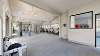 Located at 100 Riverview Pl in Jonesboro, GA, this single-floor fitness center offers a well-equipped and thoughtfully designed workout facility. The space is organized into distinct zones, providing dedicated areas for cardiovascular training, strength work, and open-floor exercise. Vaulted ceilings lend an airy, expansive feel throughout the main workout area, which is finished with neutral-toned carpet and bright white walls that contribute to a clean and motivating environment. Ceiling fans help maintain comfortable airflow during workouts.

The cardio zone is lined with large windows that frame outdoor views of surrounding greenery, filling the space with abundant natural light. Equipment includes treadmills, an elliptical trainer, and a stationary bike from TRUE Fitness. Full-length mirrors along one wall visually expand the space and support proper form during exercise. A separate area houses a cable strength machine, a dumbbell rack, and additional resistance equipment, offering a comprehensive strength training setup. An adjoining room features large grid-pane windows on multiple walls, creating a bright and open space suitable for stretching or group exercise. A storage nook holds medicine balls and accessories. The facility is well-maintained and designed to support a full range of fitness activities in a comfortable, accessible setting.