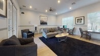 This single-floor space is designed as a comfortable and stylish lounge or common gathering area, characterized by an open, well-proportioned layout that accommodates multiple functional zones. The interior is finished with light hardwood-style flooring throughout, crisp white walls accented by a mid-height chair rail, and recessed lighting complemented by a ceiling fan, creating a bright and welcoming ambiance.

The main seating area centers around a plush gray sofa paired with a sleek oval coffee table, anchored by a deep navy area rug. Surrounding this central arrangement are additional seating options, including dark leather armchairs and patterned accent chairs with striped throw pillows, offering a cohesive yet varied mix of furnishings. A wall-mounted flat-screen television serves as a focal point in one seating nook.

A dedicated workspace with a desk and chairs is positioned near a window with blinds, allowing natural light to filter in. Framed botanical and landscape artwork adorns the walls, adding visual interest throughout.

Tall French doors open to an outdoor patio, flooding the space with natural light and providing views of lush landscaping and a paved terrace beyond, seamlessly connecting the interior to the outdoors.