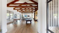 The Legacy Oak Clubhouse in Fort Worth, Texas is a thoughtfully designed community amenity space that blends modern aesthetics with warm, inviting architectural elements. The expansive single-floor layout offers generous open space throughout, anchored by a dramatic vaulted ceiling adorned with rich, exposed wood beams and skylights that flood the main hall with natural light. Wide-plank hardwood flooring runs throughout, tying each area together seamlessly.

The main recreation area features a full-size pool table and ping-pong table beneath the soaring beamed ceiling, creating an energetic social hub. Adjacent to this is a comfortable lounge area centered around a striking whitewashed stone fireplace with a mounted TV, flanked by large windows that invite ample natural light and outdoor views.

A bright dining area is outfitted with a concrete-top table, vibrant yellow chairs, and an industrial-style pendant light fixture, with oversized windows overlooking the exterior. A well-appointed kitchenette with light countertops, blue cabinetry, stainless steel appliances, and subway tile backsplash provides convenient service support. The welcoming entry features floor-to-ceiling glass doors, geometric wood wall accents, and casual seating, setting an impressive first impression for all who enter.
