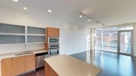 This contemporary single-floor space showcases a thoughtfully designed open-concept layout that seamlessly connects the kitchen and living areas. The kitchen is appointed with warm wood cabinetry, a large central island with a smooth light-toned countertop, and a full suite of stainless steel appliances including a built-in wall oven, microwave, and dishwasher. Open shelving adds a modern, airy touch alongside the upper cabinetry. Track lighting runs along the ceiling, providing versatile illumination throughout the space, while recessed lighting adds a clean, polished finish overhead. The living area benefits from abundant natural light streaming through expansive floor-to-ceiling windows, which frame urban views of the surrounding cityscape and provide direct access to an outdoor balcony. The polished concrete-style flooring flows continuously throughout, reinforcing the space's modern industrial aesthetic. The crisp white walls and clean architectural lines create a bright, minimalist ambiance, making this a highly functional and visually striking space suited for both everyday living and entertaining.