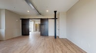 This single-floor space showcases a thoughtful blend of industrial and contemporary design elements throughout. The open living area features light wood-tone luxury vinyl plank flooring, recessed lighting, and exposed ductwork along the ceiling, lending a loft-like character to the space. A striking black structural column anchors the main area, while dark-stained sliding barn doors provide stylish transitions between rooms, adding a rustic contrast to the otherwise modern aesthetic. Large windows and a glass-paneled entry door invite natural light and outdoor views into the space.

The kitchen is well-appointed with two-tone cabinetry in gray wood-grain finishes, quartz countertops, a central island with pendant lighting, and stainless steel appliances including a gas range and French door refrigerator. In-unit stacked washer and dryer hookups are conveniently located nearby.

The bedroom features carpeted flooring, a window with outdoor views, and its own sliding barn door for privacy. Two bathrooms serve the unit: a full bath with hexagonal tile surround, a soaking tub, and matte black fixtures, and a half bath with hexagonal mosaic tile flooring, a vessel-style sink, and coordinating black hardware throughout. The overall ambiance is clean, modern, and cohesive.