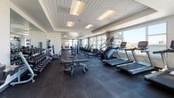 This spacious fitness center is thoughtfully designed to accommodate a comprehensive range of workout routines. The layout is open and generously proportioned, allowing ample room to move between equipment zones without feeling crowded. Large floor-to-ceiling windows line the perimeter, flooding the space with natural light and offering sweeping city skyline views that make cardio sessions particularly engaging.

The cardio zone is well-stocked with Matrix-brand treadmills, stair climbers, ellipticals, and cycling bikes, all positioned to take advantage of the panoramic views. A dedicated strength training area features a Smith machine with an adjustable bench, multiple weight benches, and a full rack of dumbbells alongside medicine balls for functional training. Cable machine stations and additional resistance equipment are distributed throughout the floor plan, offering versatility for various training styles.

Underfoot, dark rubber flooring provides both durability and cushioning across the entire space. The ceiling features a clean, modern wood-panel finish with recessed lighting that complements the natural light. The neutral color palette of white and grey walls contributes to a bright, professional atmosphere. The overall design balances functionality with a polished, upscale aesthetic.