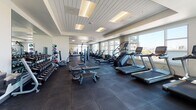 This spacious fitness center is thoughtfully designed to accommodate a comprehensive range of workout routines. The layout is open and generously proportioned, allowing ample room to move between equipment zones without feeling crowded. Large floor-to-ceiling windows line the perimeter, flooding the space with natural light and offering sweeping city skyline views that make cardio sessions particularly engaging.

The cardio zone is well-stocked with Matrix-brand treadmills, stair climbers, ellipticals, and cycling bikes, all positioned to take advantage of the panoramic views. A dedicated strength training area features a Smith machine with an adjustable bench, multiple weight benches, and a full rack of dumbbells alongside medicine balls for functional training. Cable machine stations and additional resistance equipment are distributed throughout the floor plan, offering versatility for various training styles.

Underfoot, dark rubber flooring provides both durability and cushioning across the entire space. The ceiling features a clean, modern wood-panel finish with recessed lighting that complements the natural light. The neutral color palette of white and grey walls contributes to a bright, professional atmosphere. The overall design balances functionality with a polished, upscale aesthetic.