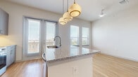Located in Northlake, TX, this well-appointed one-bedroom, one-bathroom residence offers a thoughtfully designed open-concept layout with modern finishes throughout. Light wood-tone flooring flows seamlessly across the entire space, complemented by clean white walls and vaulted ceilings that enhance the sense of openness.

The kitchen serves as the focal point of the main living area, anchored by a large granite-topped island with an undermount sink and seating. Taupe cabinetry pairs with stainless steel appliances, including a refrigerator, microwave, and range, while a set of stylish globe pendant lights adds a contemporary touch. Large windows with blinds allow abundant natural light to fill the combined kitchen and living space, which also features a ceiling fan.

A bright hallway with recessed lighting connects the main living area to the private quarters, lined with paneled doors that add architectural character. The bedroom is a generously sized, serene retreat with vaulted ceilings, a ceiling fan, and ample wall space. The bathroom features a full soaking tub with a tiled surround in soft marble tones, brushed nickel fixtures, and wood-look tile flooring. The overall ambiance is modern, clean, and move-in ready.