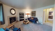 This single-floor residence offers a thoughtfully arranged layout with distinct living, dining, kitchen, bedroom, and bathroom areas. The living room serves as the heart of the home, anchored by a tile-surround fireplace and finished with soft carpeting. The space accommodates a mid-century modern aesthetic, with clean white walls that complement the warm, ambient lighting. The kitchen is functionally equipped with light maple cabinetry, ample counter space, a double-basin sink, and black appliances including a range, dishwasher, and refrigerator, all set on wood-look flooring. Adjacent to the living area, the dining room features rich hardwood-style flooring, a pendant chandelier, and large windows that invite abundant natural light and views of surrounding greenery. The bedroom is carpeted and comfortably sized, offering a double-door closet and a window for natural light. The bathroom includes a vanity with light wood cabinetry, a shower-tub combo with a curtain, and wood-look flooring. Throughout the space, neutral tones provide a versatile backdrop, while each room maintains a cohesive, modern residential character.