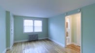 This single-floor space is characterized by its calm, inviting aesthetic, anchored by soft teal-painted walls that lend a fresh, airy quality to the interior. The main room features hardwood flooring in a light, natural finish that reflects the abundant natural light streaming through a double-window fitted with white horizontal blinds. The windows provide a bright focal point along one wall, enhancing the open feel of the room.

A classic cast-iron radiator sits beneath the windows, indicating a traditional heating system well-suited for colder climates. Clean white baseboards frame the perimeter of the room, providing a crisp contrast to the teal walls and light flooring.

An open doorway leads directly into an adjoining bathroom, visible from the main room. The bathroom features a toilet and what appears to be a mirror with wall-mounted lighting, offering convenient access and functional layout.

The overall ambiance of the space is clean, minimal, and well-lit, with a cohesive color palette of soft teal, white trim, and warm wood tones. The straightforward layout and neutral finishes make it a versatile and practical living space.