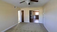 This single-story space is designed with an open, functional layout that connects the main living area to the kitchen and utility spaces. The living area is finished with wall-to-wall carpet and features a ceiling fan with a light fixture, providing both comfort and airflow. Neutral, light-toned walls throughout contribute to a clean, understated aesthetic, while white baseboards add a crisp finishing detail.

From the main living area, open doorways provide direct sightlines into both the kitchen and the laundry area. The kitchen is equipped with dark wood cabinetry, a dishwasher, and a window that allows natural light to filter in. Adjacent to the kitchen, a stacked washer and dryer unit is conveniently housed within the same open space, offering practical in-unit laundry access without requiring a separate dedicated room.

A door on the left side of the living area leads to an additional room, suggesting further private living space. The entry door on the right is solid and secure. Overall, the space offers a straightforward, efficient layout well-suited for everyday living, with all primary functional areas easily accessible from the central living zone.
