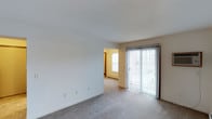 This single-floor residence features two bedrooms and one and a half bathrooms, offering a functional and comfortable layout. The living area is bright and open, highlighted by a full-length sliding glass door with vertical blinds that invites natural light and provides access to an outdoor space with pleasant exterior views. Neutral-toned carpet flooring extends throughout, adding warmth underfoot and contributing to a cozy ambiance. The walls are finished in clean white and soft warm tones, creating a light and airy feel. A baseboard heating unit is visible along the perimeter, ensuring year-round comfort. An adjacent room, visible through a connecting opening, features the same warm wall tones and an additional window that further enhances the natural light within the home. The layout connects spaces in a straightforward and accessible manner, making the floor plan easy to navigate. The half bathroom provides added convenience alongside the full bathroom, catering well to everyday living. Overall, this residence presents a simple, clean, and functional living environment suitable for a variety of lifestyles.