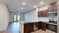 Located in Prosper, TX, this well-appointed 1-bedroom, 1-bathroom residence offers a thoughtfully designed open-concept layout that seamlessly connects the kitchen, dining, and living areas. The kitchen is a standout feature, showcasing rich dark-wood cabinetry, granite countertops, a large peninsula with a double undermount sink and gooseneck faucet, and a full suite of stainless steel appliances including a refrigerator, range, microwave, and dishwasher. Wood-style flooring flows throughout the main living areas, complemented by crown molding and recessed lighting that lend a polished finish. The living room is bright and airy, anchored by a ceiling fan and French doors that invite natural light and provide outdoor access. The spacious bedroom is finished with plush carpet, a ceiling fan, and a generous window for added natural light, supported by two closets for ample storage. The bathroom continues the design consistency with granite countertops, dark cabinetry, and wood-style flooring. A dedicated laundry area adds everyday convenience. Neutral tones throughout create a clean, modern ambiance well-suited to a variety of interior styles.