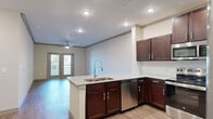 Located in Prosper, TX, this well-appointed 1-bedroom, 1-bathroom residence offers a thoughtfully designed open-concept layout that seamlessly connects the kitchen, dining, and living areas. The kitchen is a standout feature, showcasing rich dark-wood cabinetry, granite countertops, a large peninsula with a double undermount sink and gooseneck faucet, and a full suite of stainless steel appliances including a refrigerator, range, microwave, and dishwasher. Wood-style flooring flows throughout the main living areas, complemented by crown molding and recessed lighting that lend a polished finish. The living room is bright and airy, anchored by a ceiling fan and French doors that invite natural light and provide outdoor access. The spacious bedroom is finished with plush carpet, a ceiling fan, and a generous window for added natural light, supported by two closets for ample storage. The bathroom continues the design consistency with granite countertops, dark cabinetry, and wood-style flooring. A dedicated laundry area adds everyday convenience. Neutral tones throughout create a clean, modern ambiance well-suited to a variety of interior styles.