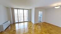 This single-floor space is characterized by a clean, contemporary layout with an open and airy feel. The main living area showcases polished parquet flooring arranged in a classic checkerboard pattern, lending warmth and a timeless aesthetic to the room. Large floor-to-ceiling sliding glass doors flood the space with abundant natural light and open directly onto a private balcony, offering views of the surrounding urban skyline. The balcony features a simple railing and provides an outdoor retreat connected seamlessly to the interior.

The living area transitions naturally into a separate kitchen, accessible through a dedicated doorway. The kitchen is equipped with white cabinetry, a gas range, and a built-in microwave, presenting a functional and well-organized cooking space. The white walls and neutral tones throughout the unit create a bright, welcoming atmosphere that complements the natural light streaming in from the balcony. The overall layout is efficient and thoughtfully designed, making strong use of the available space while maintaining an open, uncluttered feel throughout.
