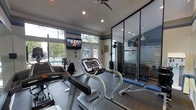 This single-floor fitness center is thoughtfully designed to provide a comprehensive workout environment in a compact yet functional space. The room is outfitted with multiple treadmills and a stationary bike for cardiovascular training, complemented by a cable strength machine and a weight bench for resistance workouts. Recessed lighting throughout the ceiling ensures the space is well-lit, while a ceiling fan promotes air circulation during intense exercise sessions. A wall-mounted flat-screen television adds entertainment value for those looking to stay engaged during cardio workouts. One of the standout features is the full-length mirrored wall partition, which visually expands the room and allows users to monitor their form during exercise. The flooring consists of durable rubber gym matting, providing both safety and comfort underfoot. Natural light flows in through a set of windows and glass-paneled doors that face an outdoor pool area, creating an open and refreshing ambiance. The walls are accented with a neutral color palette offset by a dark decorative band near the ceiling, giving the space a clean, modern aesthetic. Overall, this fitness center strikes a balance between functionality and comfort, offering residents or users a well-rounded exercise facility.
