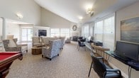 This single-floor clubhouse offers a well-appointed communal space designed to accommodate a variety of activities and social gatherings. The main lounge area is immediately striking, featuring soaring vaulted ceilings that lend an open, airy ambiance throughout. Neutral tones on the walls and plush carpeted flooring create a warm, welcoming atmosphere, while abundant natural light streams in through multiple large windows dressed with white plantation shutters.

The lounge is comfortably furnished with an eclectic mix of patterned accent chairs, a generously sized sofa with colorful throw pillows, and rustic wooden side tables, all centered around a decorative fireplace that serves as a focal point. A dedicated billiards area with a full-sized pool table adds a recreational dimension to the space.

A built-in business center along one wall provides computer workstations and a printer, catering to residents' professional needs. The kitchen area is thoughtfully outfitted with white cabinetry, granite countertops, stainless steel appliances, and a breakfast bar with woven rattan stools, making it ideal for community events and casual dining. Dark hardwood-style flooring in the kitchen contrasts beautifully with the carpeted lounge, clearly defining each functional zone within this versatile clubhouse.