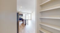 This well-designed single-floor unit showcases a contemporary open-concept layout that seamlessly connects the kitchen, dining, and living areas. The kitchen is a standout feature, equipped with a full suite of stainless steel appliances — including a refrigerator, range, microwave, and dishwasher — complemented by rich dark wood cabinetry, a classic white tile backsplash, and granite countertops. A central island with a granite surface and bar-height legs provides additional prep space and casual dining. Pendant lighting above the island and recessed ceiling fixtures throughout ensure bright, well-lit spaces. Light-toned hardwood flooring runs continuously across the unit, contributing to a clean, cohesive aesthetic. The living area is generously proportioned, featuring a ceiling fan and large windows with blinds that invite ample natural light. Built-in open shelving near the entryway offers practical display and storage options. The bedroom is bright and airy with a ceiling fan, a large window, and access to a walk-in closet with built-in shelving. The bathroom features a dark wood vanity with granite countertops, a framed mirror, above-vanity cabinet storage, and a full tub-shower combination. Overall, the unit presents a modern, functional, and well-appointed living environment.