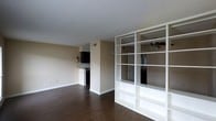 This single-floor space features an open-concept layout that connects the living area to the kitchen, creating a functional and flexible floor plan. A standout architectural element is the large, built-in white open shelving unit, which serves as a room divider while maintaining visual openness between spaces. The shelving offers generous storage and display potential without closing off the flow of the interior.

The flooring throughout is a rich, dark hardwood, adding warmth and contrast against the neutral beige walls and crisp white trim. A ceiling fan is visible beyond the divider, providing both air circulation and a functional lighting fixture. The kitchen area, positioned toward the rear, features dark cabinetry and a countertop peninsula, offering a natural transition between cooking and living areas.

The walls are painted in a soft, neutral tone that keeps the space feeling bright and adaptable to various décor styles. White baseboards run cleanly along the perimeter, complementing the overall clean aesthetic. The open layout and thoughtful use of the built-in divider make this space well-suited for both relaxed living and practical everyday use.